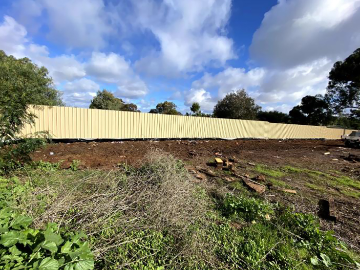 Overgrown block being cleared, with a long cream steel fence behind