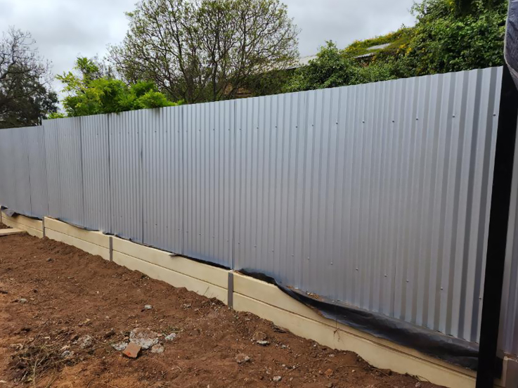 Grey corrugated steel fence on a sleeper base along a garden bed